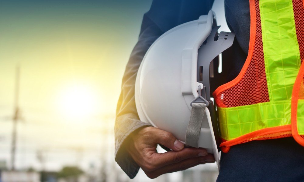 A safety technician stands on a jobsite holding a white hard hat and wearing a bright orange vest with a yellow stripe.
