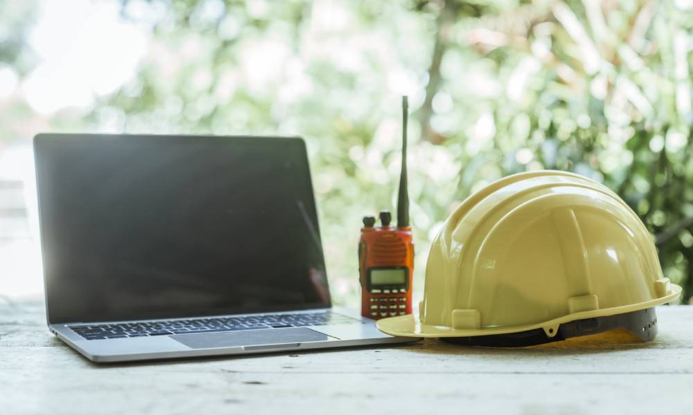 An open laptop, a yellow hardhat, and an orange walkie talkie all sit lined up on an wooden desk surface.