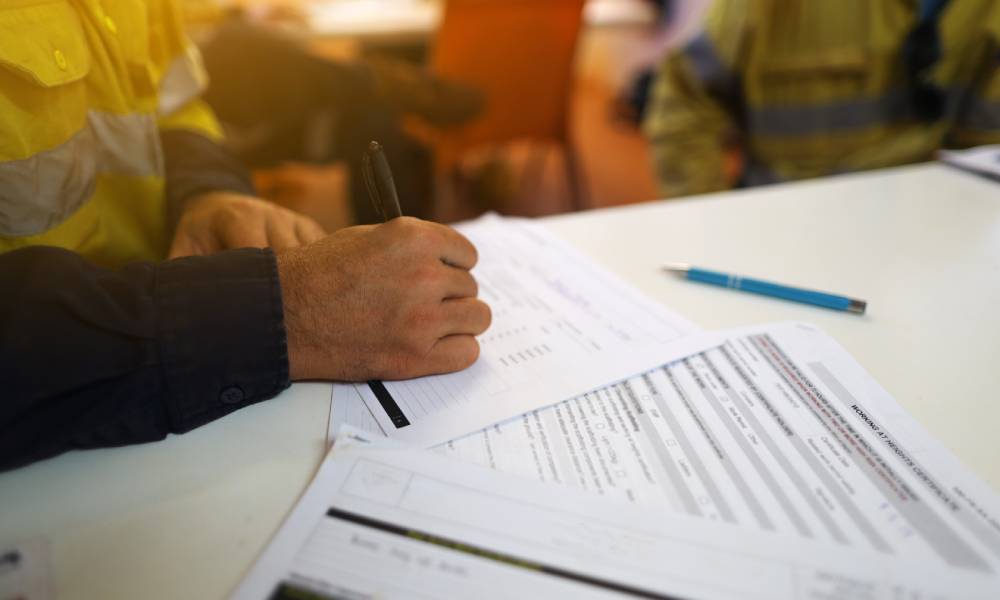 A person in a black shirt and bright yellow vest is writing on a piece of paper on a desk surrounded by other papers.