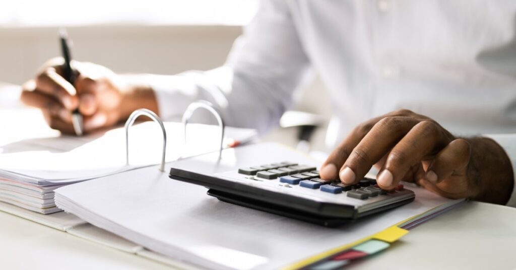 A business person sits at a desk going through a binder full of papers. They're crunching numbers on a calculator.