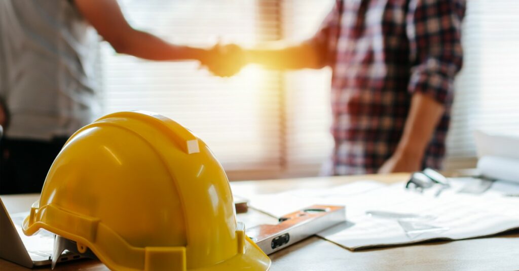 A yellow hard hat sits on a desk with a level and construction plans. Two contractors shake hands in the background.