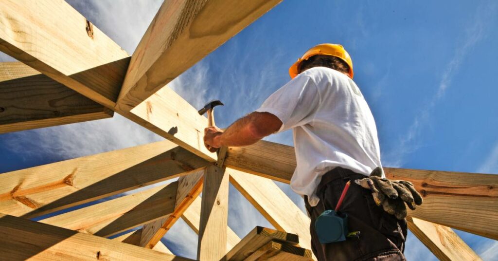 A construction worker is working on hammering in a nail. They are wearing a yellow hard hat and a white shirt.