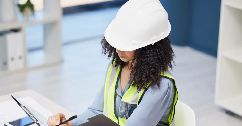 A woman wearing a white hard hat and high-visibility vest sitting at a desk while writing on a document.