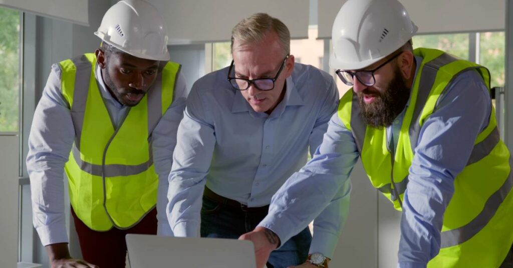 A man standing at a desk looking at an open laptop. Two contractors wearing hard hats and high-visibility vests flank him.