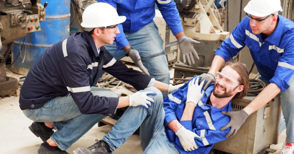 A worker wearing a blue shirt on a manufacturing floor lying down holding his shoulder in pain surrounded by other workers.