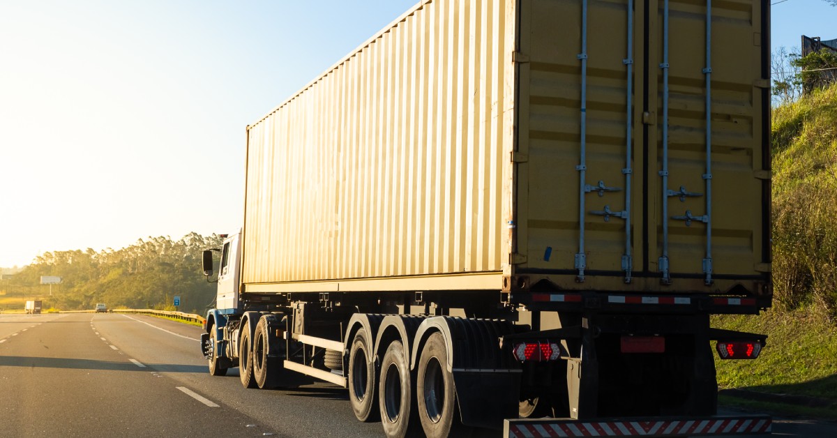 A semi truck driving in the right lane of a highway. Its brake lights are on and there are reflectors on the back of the truck.