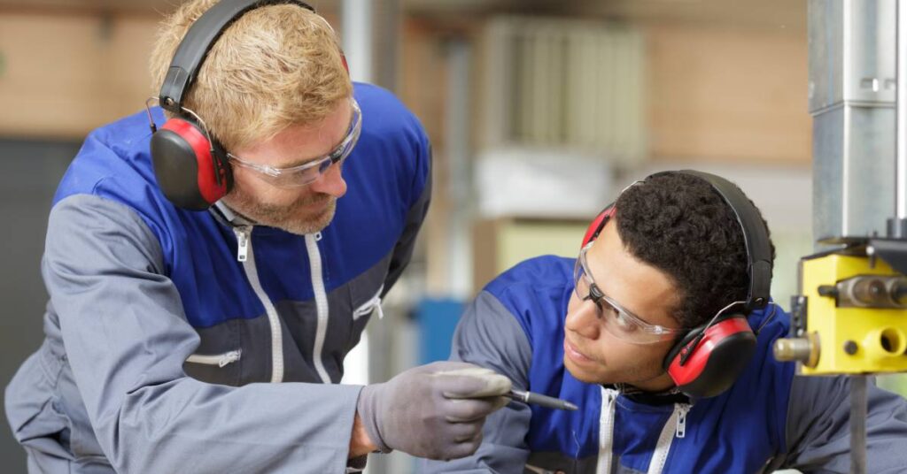 A worker in an industrial setting standing next to another person at a machine training them to use it properly.