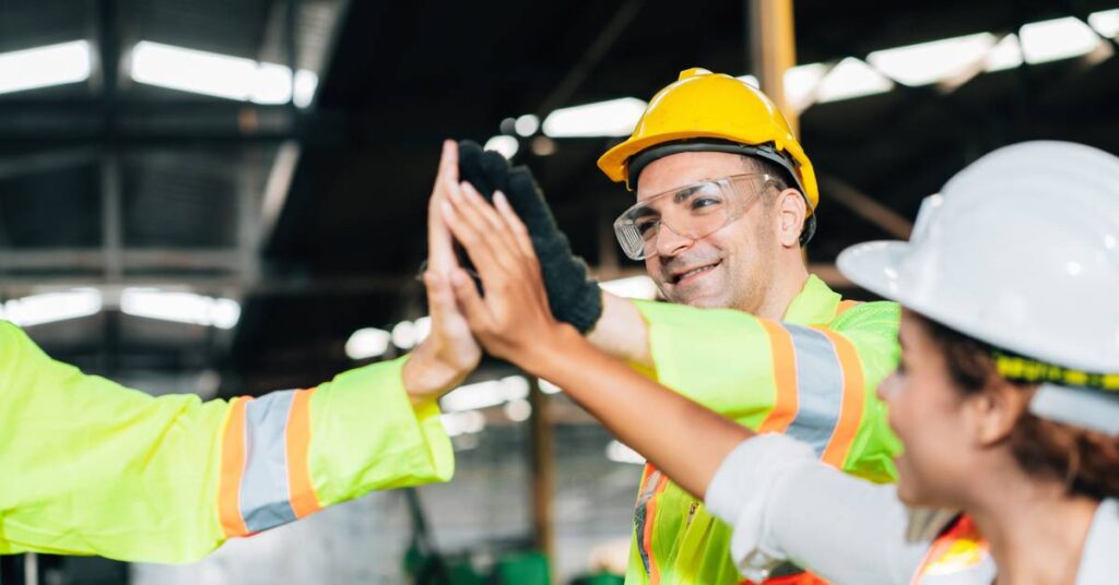 Three workers in an industrial setting wearing high-visibility gear holding their hands out to give one another high-fives.