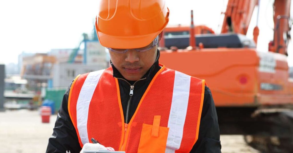 A worker wearing an orange hard hat and an orange and white vest writing on a clipboard. A forklift is in the background.