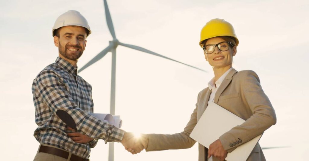 A man and a woman wearing professional clothing and hardhats while smiling and shaking hands in front of a wind turbine.