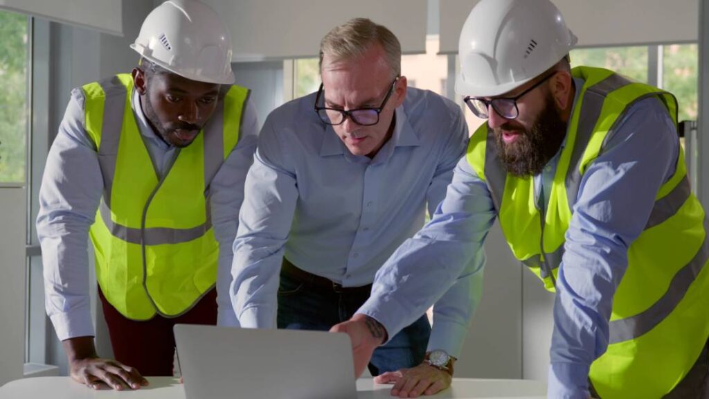 A man wearing a blue button-down shirt speaks to two men wearing yellow safety vests. They're looking and pointing at a laptop.