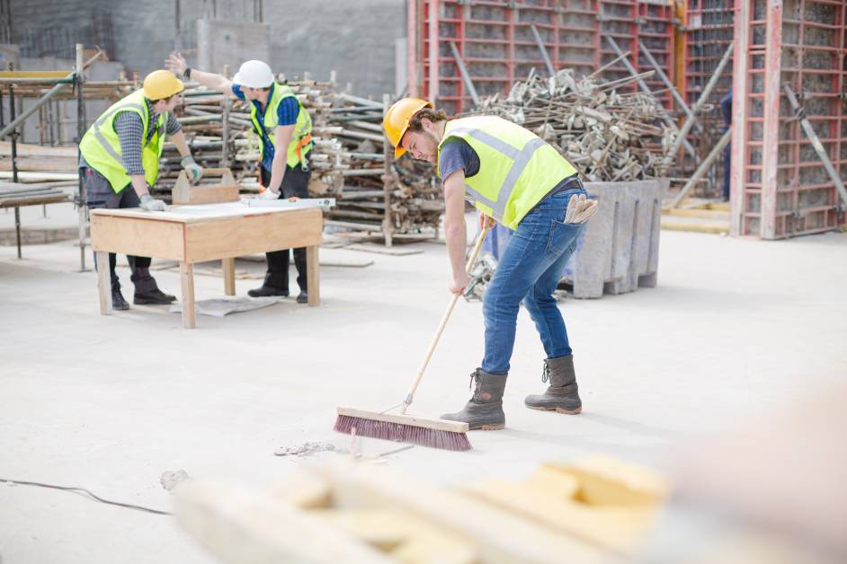 A man wearing a yellow safety vest and hard hat sweeps dust and debris. Two workers are talking in the background.